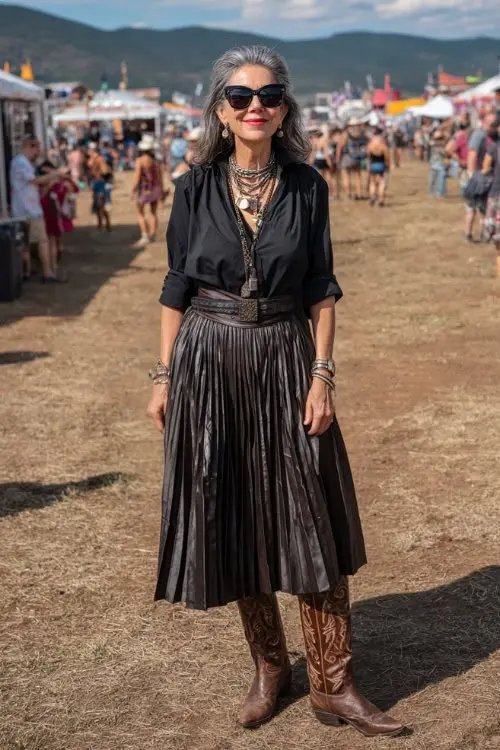 Woman over 50 in a black pleated midi skirt and black top with tall brown cowboy boots