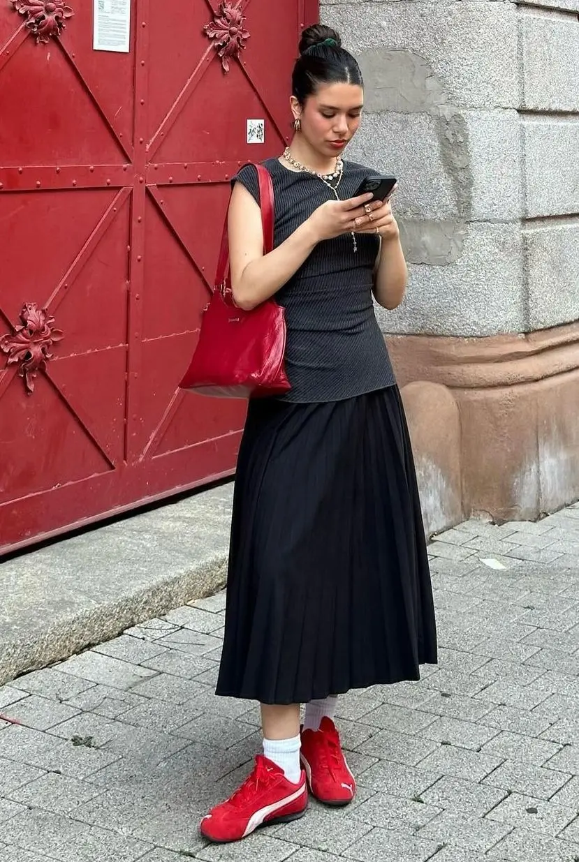 Woman wearing a black pleated dress with a red bag and red sneakers beside a red door