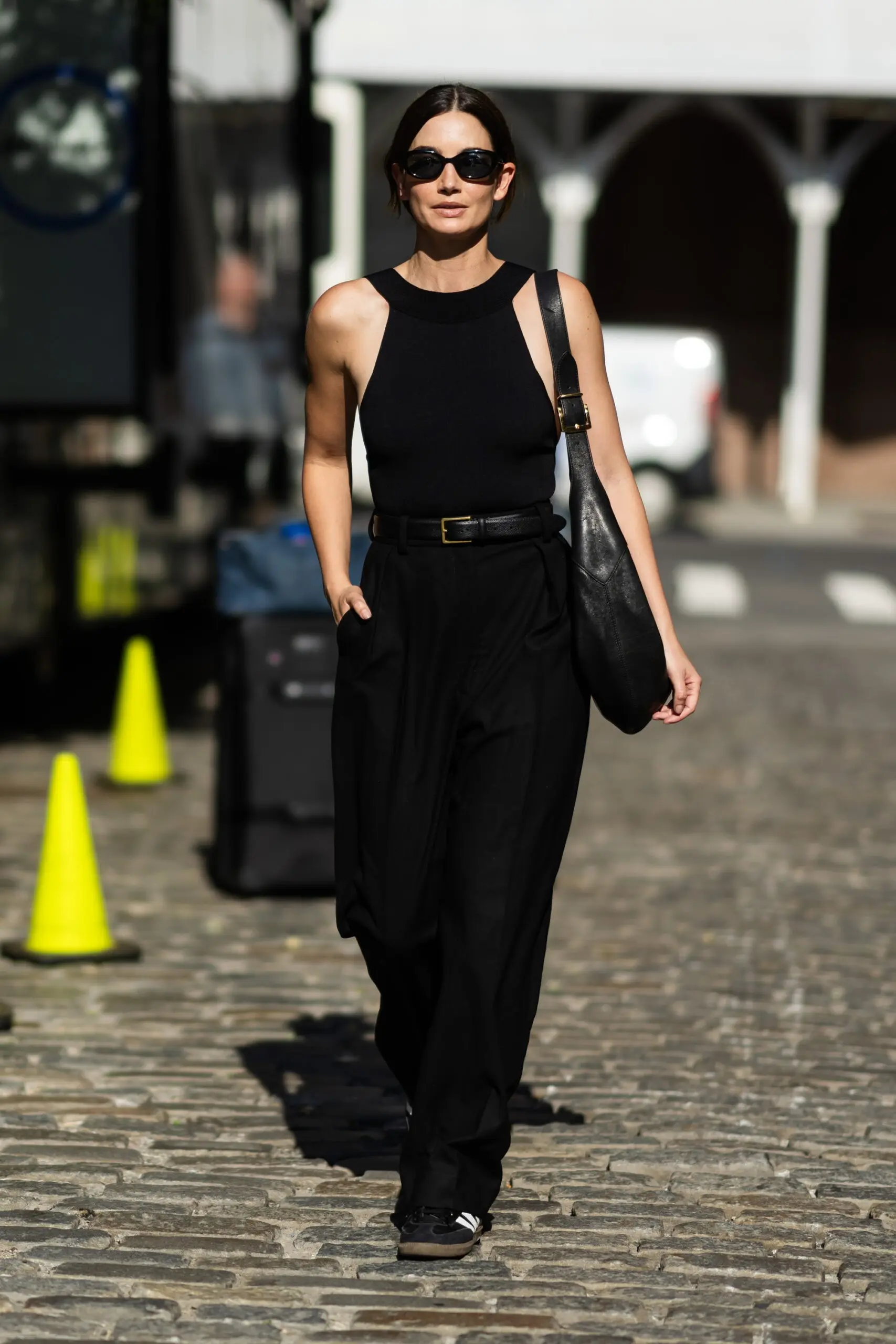 Woman wearing a black sleeveless bodysuit top with black wide-leg trousers and sunglasses on a cobblestone street